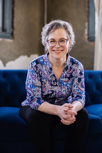 Christa Wessel woman with glasses sitting on a blue couch, leaning slightly forward