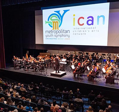 A youth orchestra sitting under a brightly lit screen